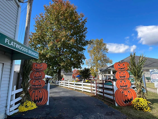 Entrance to a fall festival with large wooden pumpkin cutouts stacked on each side of a paved path, surrounded by autumn trees and white fences.