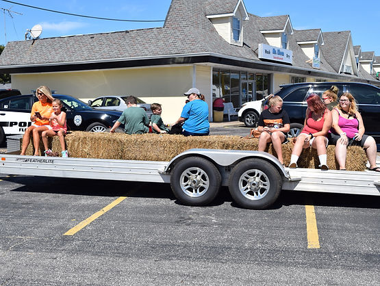 People sit on hay bales arranged on a flatbed trailer in a parking lot, preparing for a hayride on a sunny day.