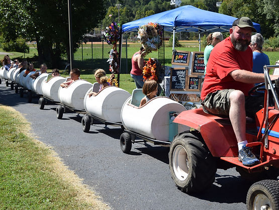 A man drives a small red tractor pulling a train of white barrel cars filled with children at an outdoor fair. A vendor tent and decorated wreaths are visible in the background.