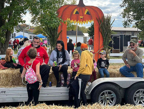 Families and children sit on hay bales on a decorated trailer featuring a large wooden pumpkin cutout with a carved face at an outdoor fall festival.